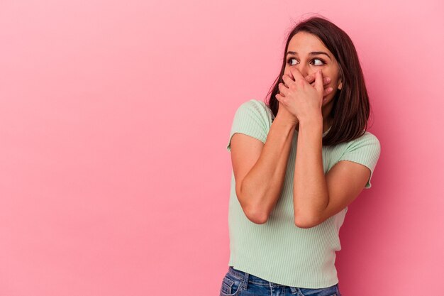 Giovane donna caucasica isolata su sfondo rosa premurosa in cerca di uno spazio copia che copre la bocca con la mano.