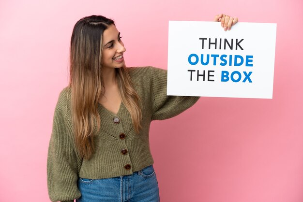 Giovane donna caucasica isolata su sfondo rosa, tenendo un cartello con il testo Think Outside The Box