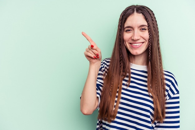 Giovane donna caucasica isolata su sfondo verde sorridente allegramente indicando con l'indice lontano