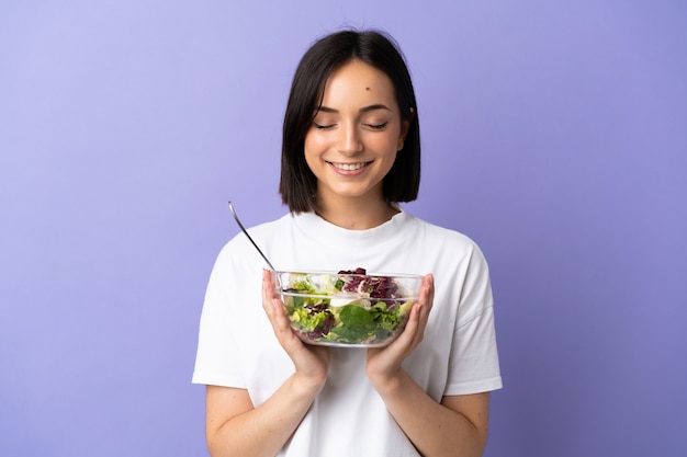 Giovane donna caucasica isolata su sfondo viola con in mano una ciotola di insalata con espressione felice