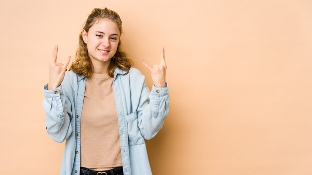 Giovane donna caucasica isolata sul beige che mostra un gesto di corna come un concetto di rivoluzione.