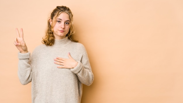 Giovane donna caucasica isolata sul muro beige prestando giuramento, mettendo la mano sul petto.