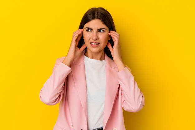 Giovane donna caucasica isolata sul muro giallo che copre le orecchie con le dita, stressata e disperata da un ambiente rumoroso.