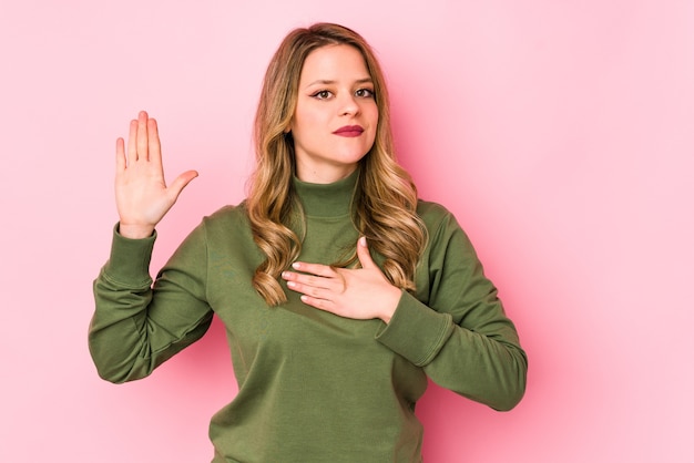 Giovane donna caucasica isolata sul muro rosa prestando giuramento, mettendo la mano sul petto.