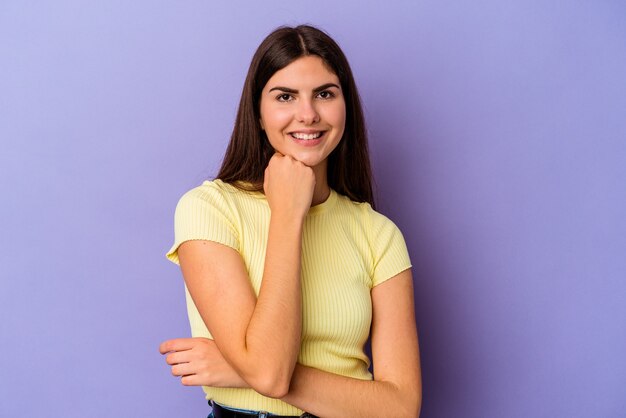 Giovane donna caucasica isolata sul muro viola sorridente felice e fiduciosa, toccando il mento con la mano