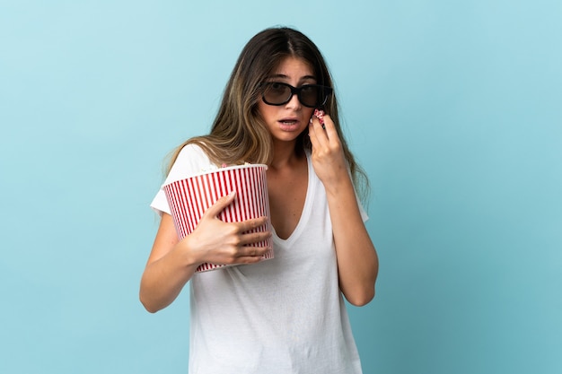 Giovane donna caucasica isolata sull'azzurro con gli occhiali 3d e che tiene un grande secchio di popcorn