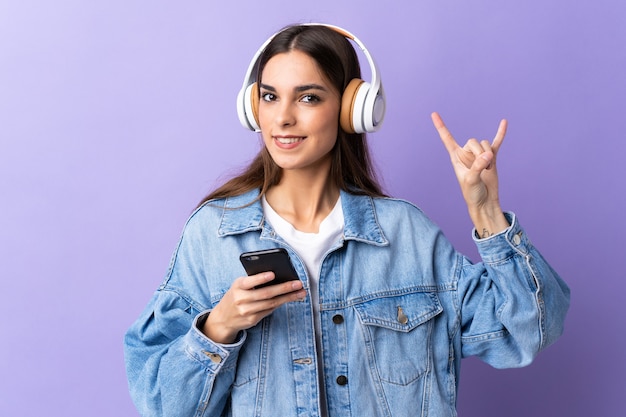 Giovane donna caucasica isolata sulla musica d'ascolto del fondo viola con un gesto di roccia che fa mobile