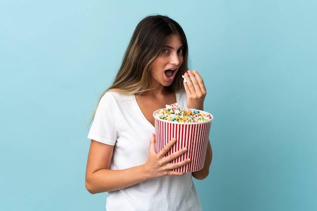 Giovane donna caucasica isolata sulla parete blu che tiene un grande secchio di popcorn
