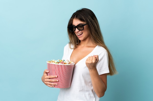 Giovane donna caucasica isolata sulla parete blu con gli occhiali 3d e che tiene un grande secchio di popcorn mentre guarda di lato