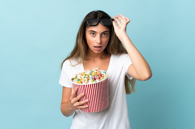 Giovane donna caucasica isolata sulla parete blu sorpresa con gli occhiali 3d e che tiene un grande secchio di popcorn