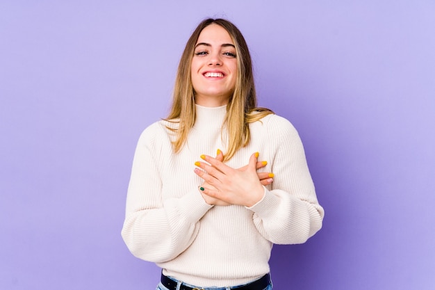 Giovane donna caucasica isolata sulla parete viola che ride mantenendo le mani sul cuore, il concetto di felicità.