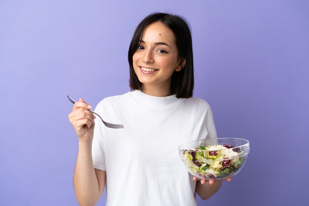 Giovane donna caucasica isolata sulla parete viola che tiene una ciotola di insalata con l'espressione felice