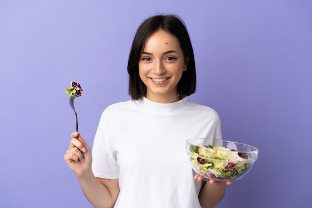 Giovane donna caucasica isolata sulla porpora che tiene una ciotola di insalata con l'espressione felice