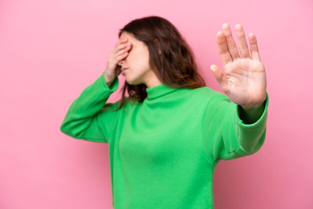 Giovane donna caucasica isolata sullo sfondo rosa che fa il gesto di fermarsi e copre la faccia