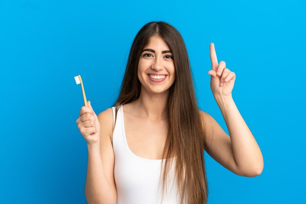 Giovane donna caucasica lavarsi i denti isolati su sfondo blu che punta verso l'alto una grande idea