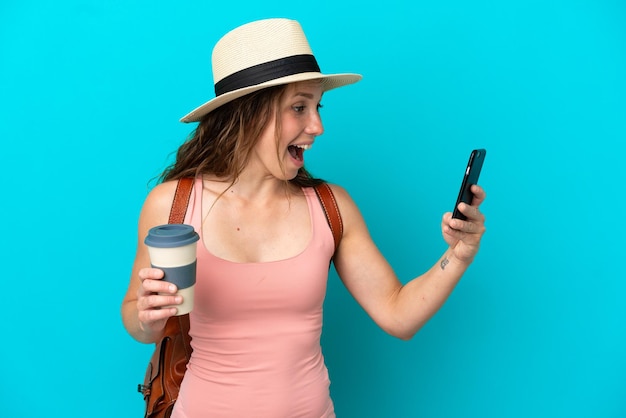Giovane donna caucasica nelle vacanze estive isolata su sfondo blu che tiene il caffè da portare via e un cellulare