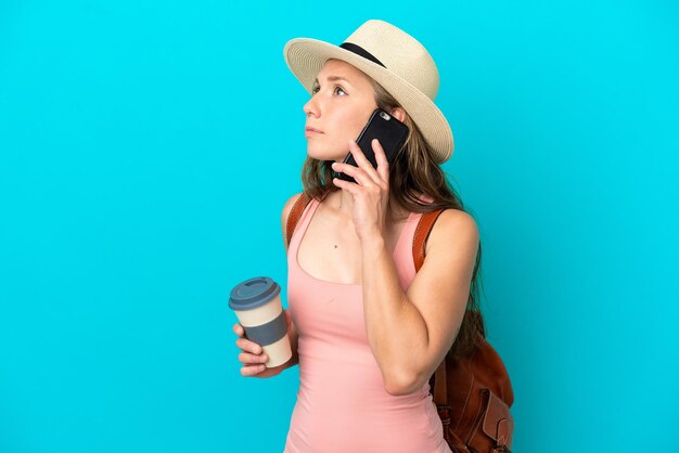 Giovane donna caucasica nelle vacanze estive isolata su sfondo blu che tiene il caffè da portare via e un cellulare