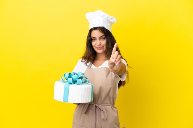 Giovane donna caucasica pasticcere con una grande torta isolata su sfondo giallo che mostra e solleva un dito