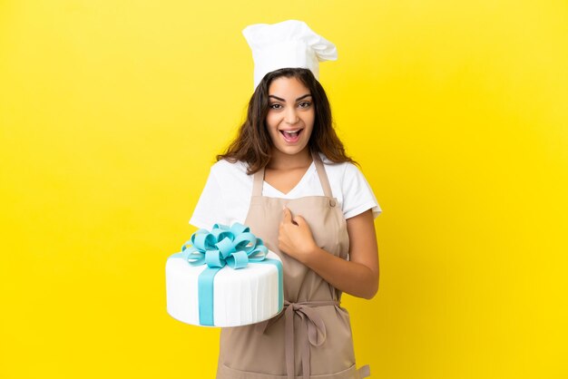 Giovane donna caucasica pasticcere con una grande torta isolata su sfondo giallo con espressione facciale a sorpresa