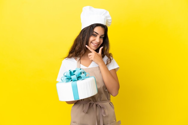 Giovane donna caucasica pasticcere con una grande torta isolata su sfondo giallo felice e sorridente