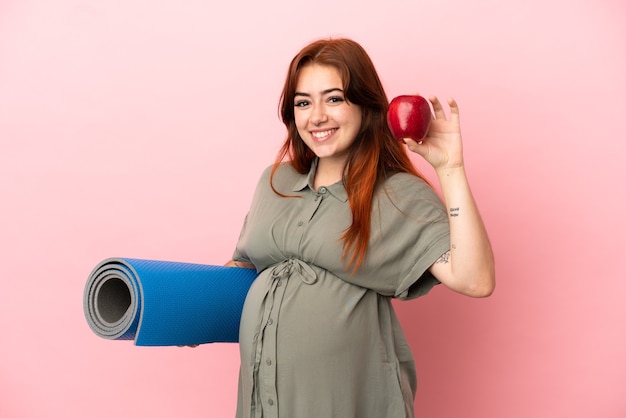 Giovane donna caucasica rossa isolata su sfondo rosa incinta che tiene una mela e va a lezioni di yoga