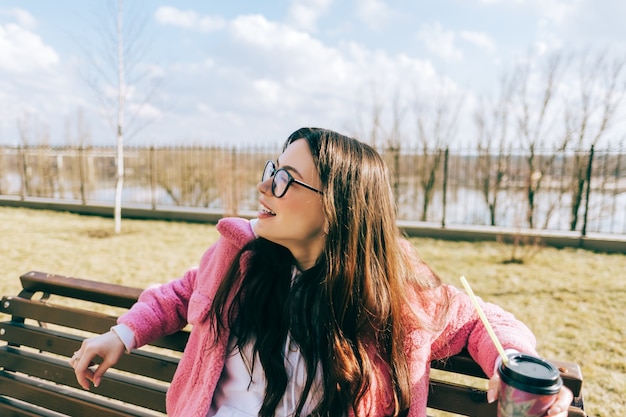 Giovane donna caucasica seduta su una panchina nel parco e bere caffè.