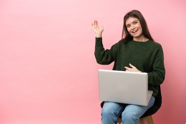 Giovane donna caucasica seduta su una sedia con il suo computer portatile isolato su sfondo rosa che fa il gesto della chitarra