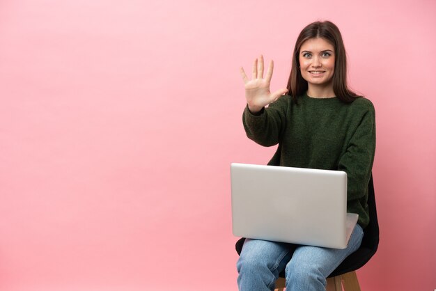 Giovane donna caucasica seduta su una sedia con il suo computer portatile isolato su sfondo rosa contando cinque con le dita