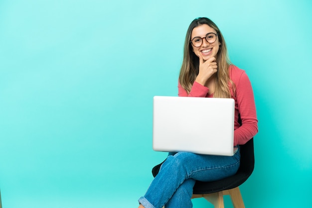 Giovane donna caucasica seduta su una sedia con il suo pc isolato su sfondo blu con gli occhiali e sorridente