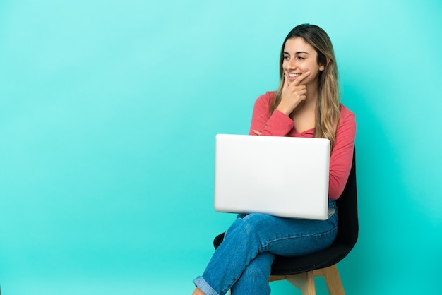 Giovane donna caucasica seduta su una sedia con il suo pc isolato su sfondo blu guardando di lato