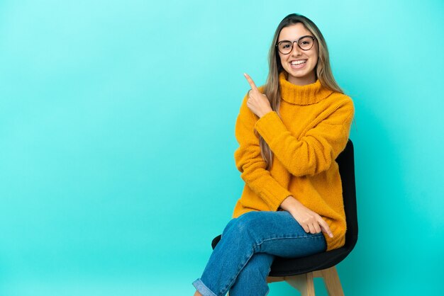 Giovane donna caucasica seduta su una sedia isolata su sfondo blu che punta di lato per presentare un prodotto