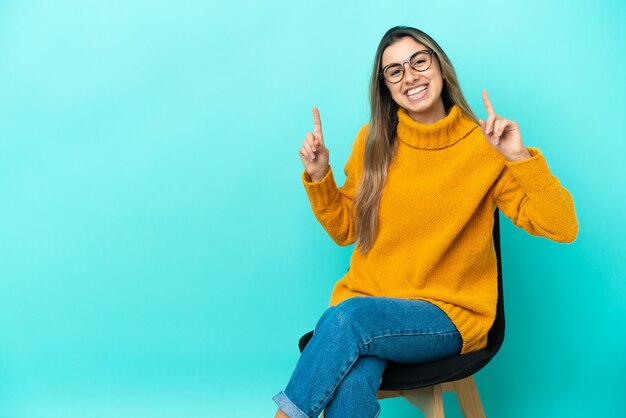 Giovane donna caucasica seduta su una sedia isolata su sfondo blu rivolto verso l'alto una grande idea