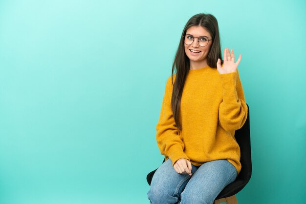 Giovane donna caucasica seduta su una sedia isolata su sfondo blu salutando con la mano con espressione felice