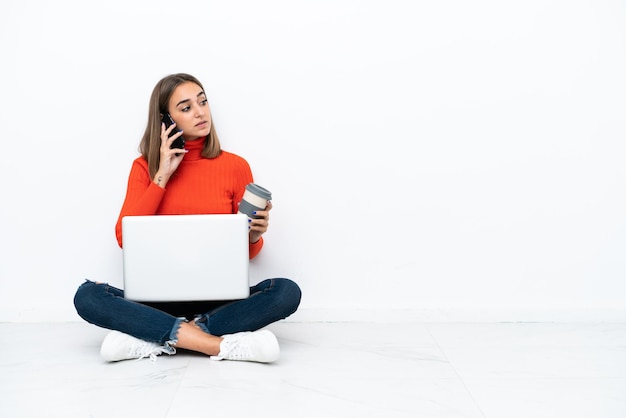 Giovane donna caucasica seduta sul pavimento con un computer portatile che tiene il caffè da portare via e un cellulare