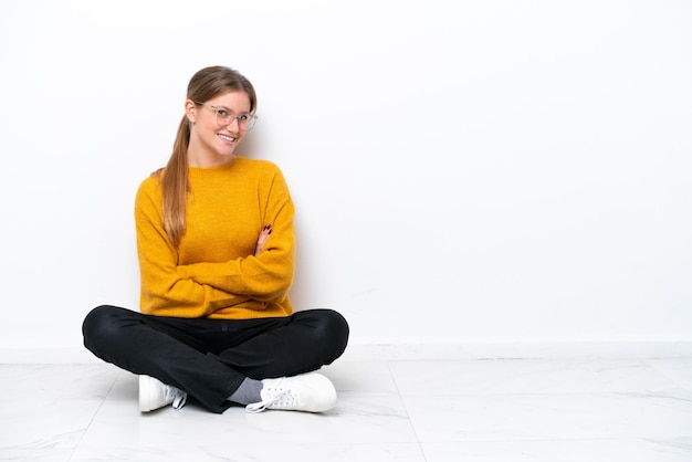 Giovane donna caucasica seduta sul pavimento isolata su sfondo bianco con le braccia incrociate e guardando avanti