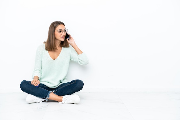 Giovane donna caucasica seduta sul pavimento isolata su sfondo bianco mantenendo una conversazione con il telefono cellulare