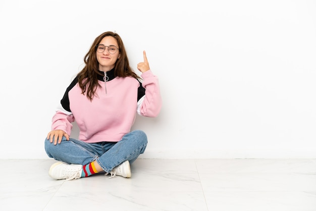 Giovane donna caucasica seduta sul pavimento isolato su sfondo bianco che indica con il dito indice una grande idea