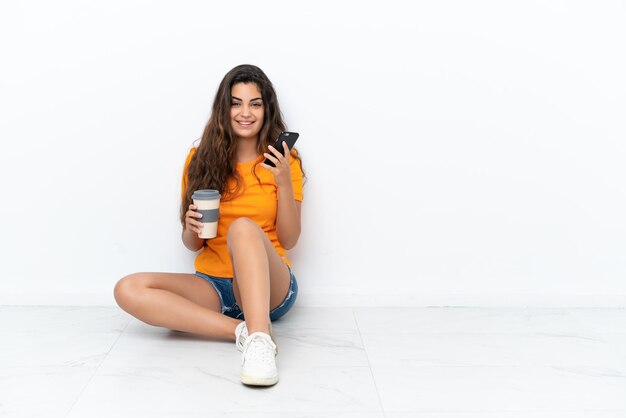 Giovane donna caucasica seduta sul pavimento isolato su sfondo bianco con in mano caffè da asporto e un cellulare