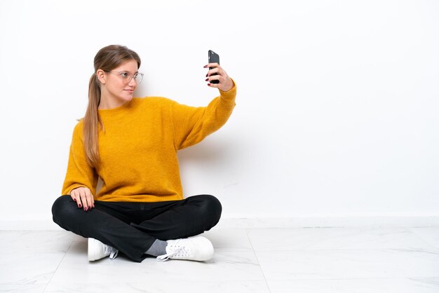 Giovane donna caucasica seduta sul pavimento isolato su sfondo bianco facendo un selfie