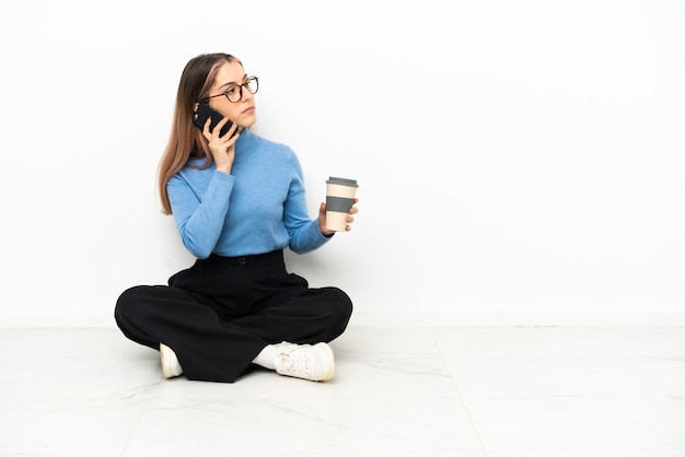 Giovane donna caucasica seduta sul pavimento tenendo il caffè da portare via e un cellulare