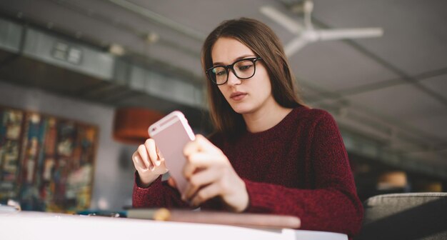 Giovane donna caucasica seria con gli occhiali che controlla il saldo bancario tramite app sul telefono