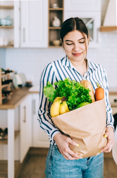 Giovane donna caucasica sorridente tenere eco shopping bag con verdure fresche e baguette in cucina moderna.