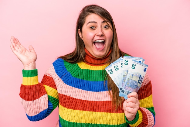 Giovane donna caucasica sovrappeso che tiene banconote isolate su sfondo rosa ricevendo una piacevole sorpresa, eccitata e alzando le mani.