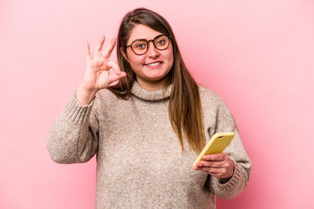 Giovane donna caucasica sovrappeso in possesso di un telefono cellulare isolato su sfondo rosa allegra e sicura di sé che mostra un gesto ok.