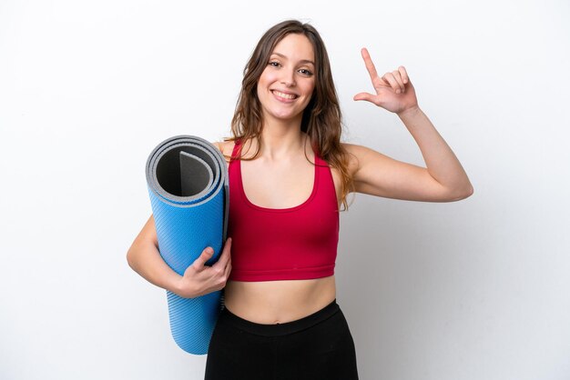 Giovane donna caucasica sportiva che va a lezioni di yoga mentre tiene un tappetino isolato su sfondo bianco che indica una grande idea