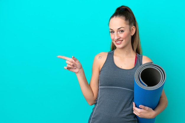 Giovane donna caucasica sportiva che va a lezioni di yoga mentre tiene un tappetino isolato su sfondo blu che punta il dito di lato