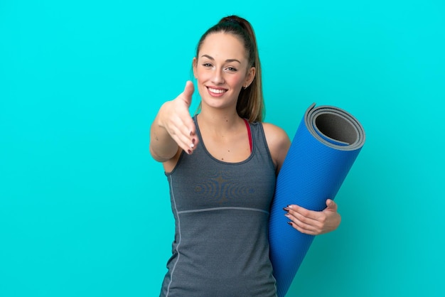 Giovane donna caucasica sportiva che va a lezioni di yoga mentre tiene un tappetino isolato su sfondo blu che stringe la mano per chiudere un buon affare