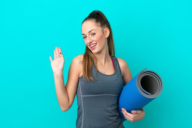 Giovane donna caucasica sportiva che va a lezioni di yoga mentre tiene un tappetino isolato su sfondo blu salutando con la mano con espressione felice