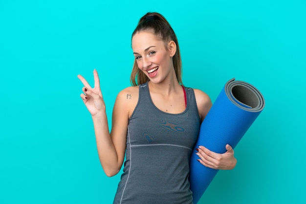 Giovane donna caucasica sportiva che va a lezioni di yoga mentre tiene un tappetino isolato su sfondo blu sorridente e mostra il segno di vittoria