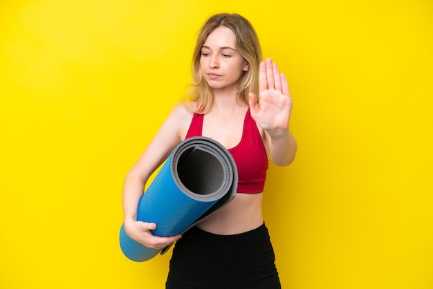 Giovane donna caucasica sportiva che va alle lezioni di yoga mentre tiene un tappetino isolato su sfondo giallo facendo gesto di arresto e deluso